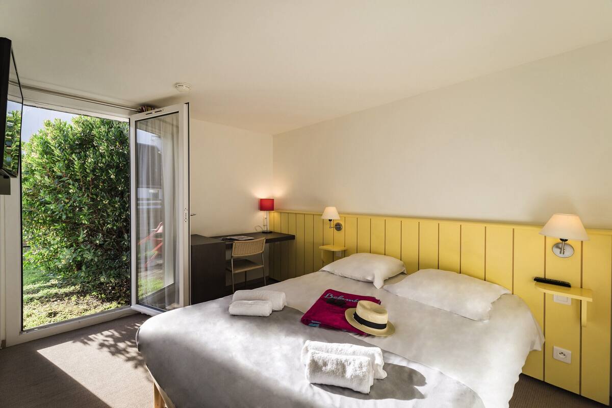A hotel room with a bed, a desk, and a chair. There is a window with a view of greenery outside.