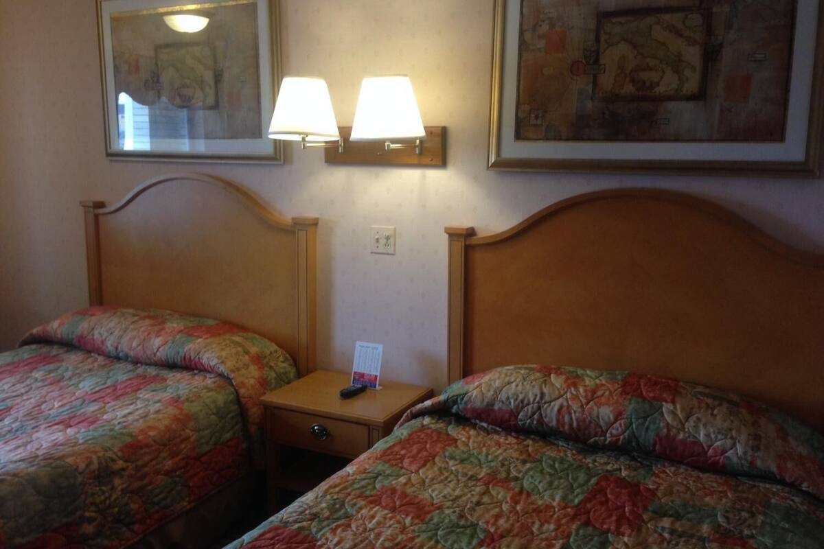 A hotel room with two single beds, each with a floral-patterned bedspread. There are two wall-mounted lamps above the beds, and a small table between them.