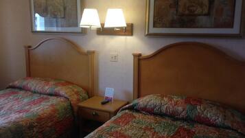 A hotel room with two single beds, each with a floral-patterned bedspread. There are two wall-mounted lamps above the beds, and a small table between them.