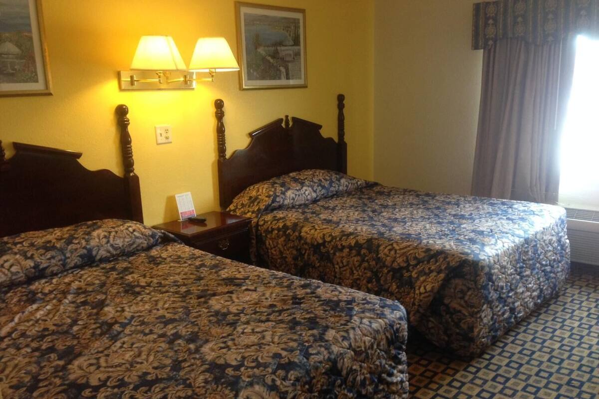 A hotel room with two beds, a nightstand, a lamp, and a window with curtains.