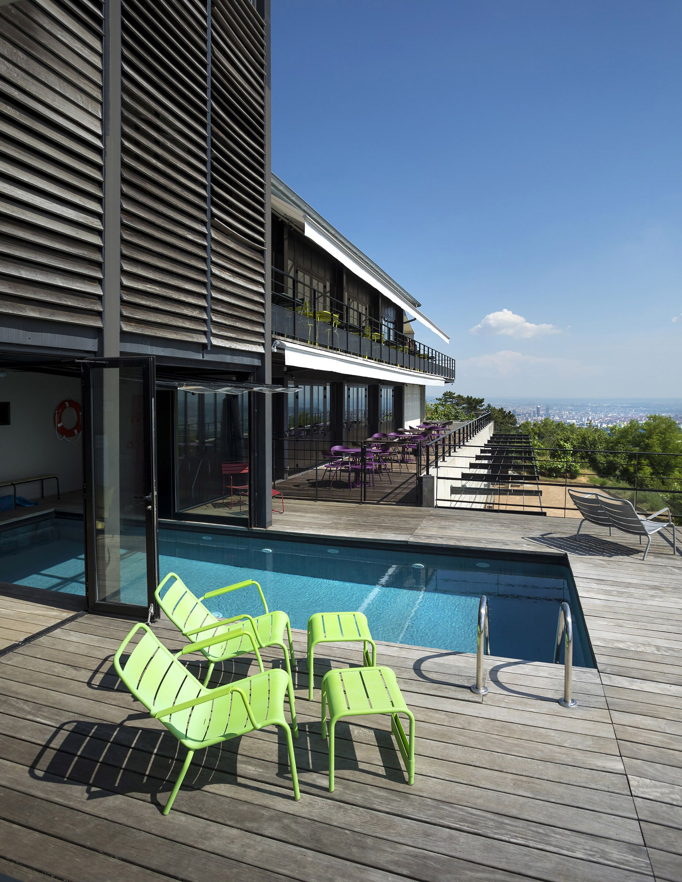 A modern building with a pool area featuring green chairs and a view of the city.