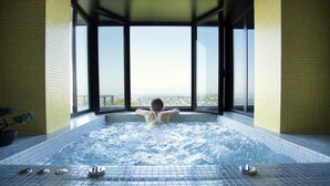 A person relaxing in a hot tub with a city view through large windows. - L'Ermitage Hôtel Cuisine-à-Manger (Saint-Cyr-au-Mont-d'Or)