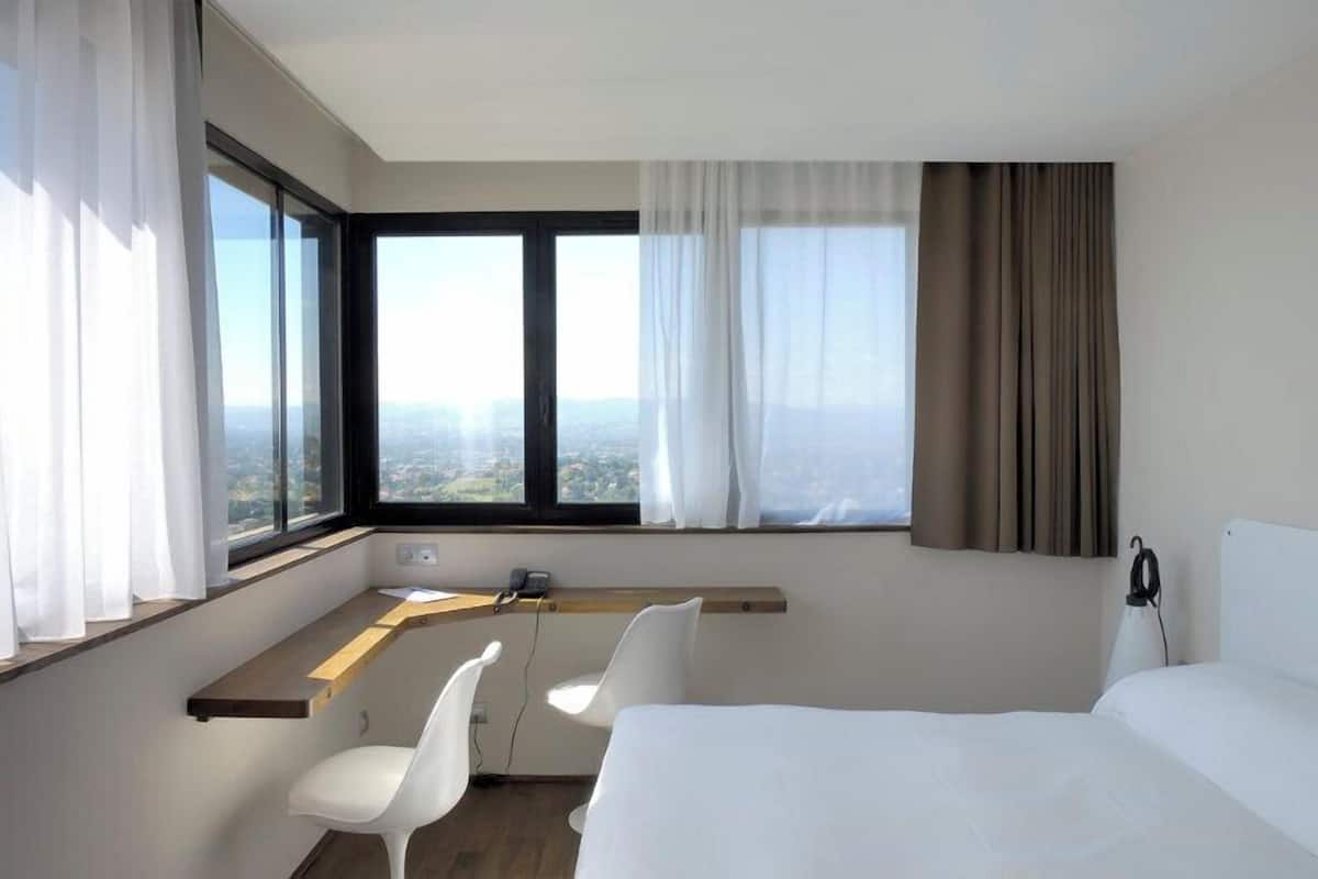 A hotel room with a bed, a desk with a chair, a telephone, and a window with curtains.