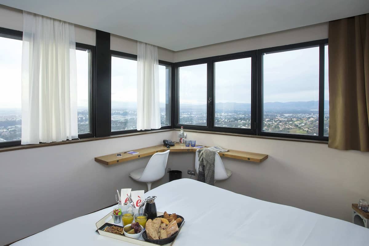 A hotel room with a large bed, a desk with a chair, and a view of a cityscape through large windows.