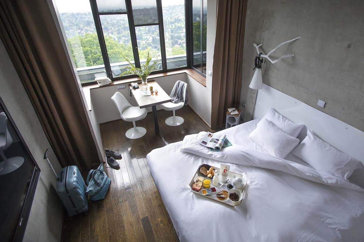 A hotel room with a bed, breakfast tray, and a view of a cityscape through large windows.