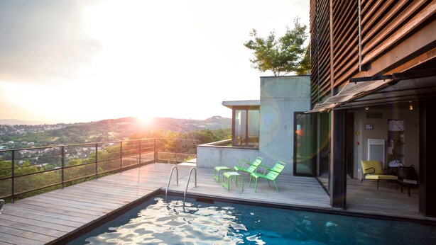 A modern outdoor pool area with a wooden deck, green chairs, and a view of a cityscape and hills in the background. - L'Ermitage Hôtel Cuisine-à-Manger (Saint-Cyr-au-Mont-d'Or)