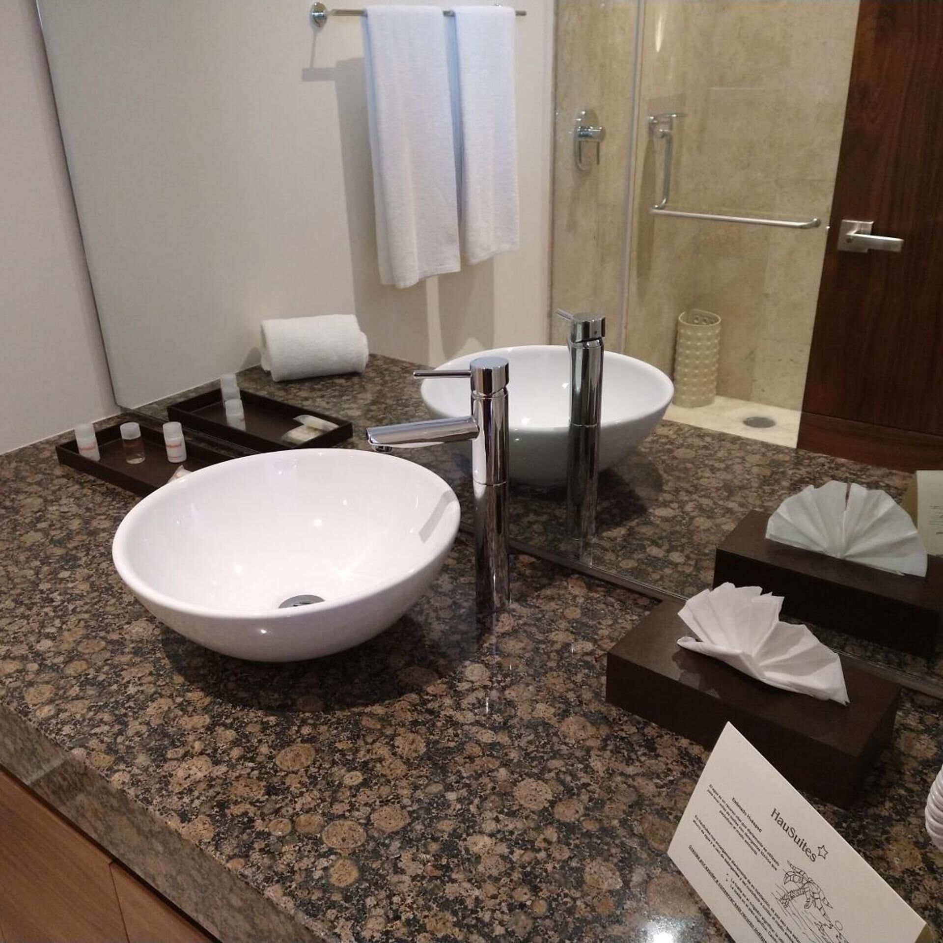 A bathroom with a white sink, a mirror, a towel rack, a shower, and a sign indicating the hotel name.