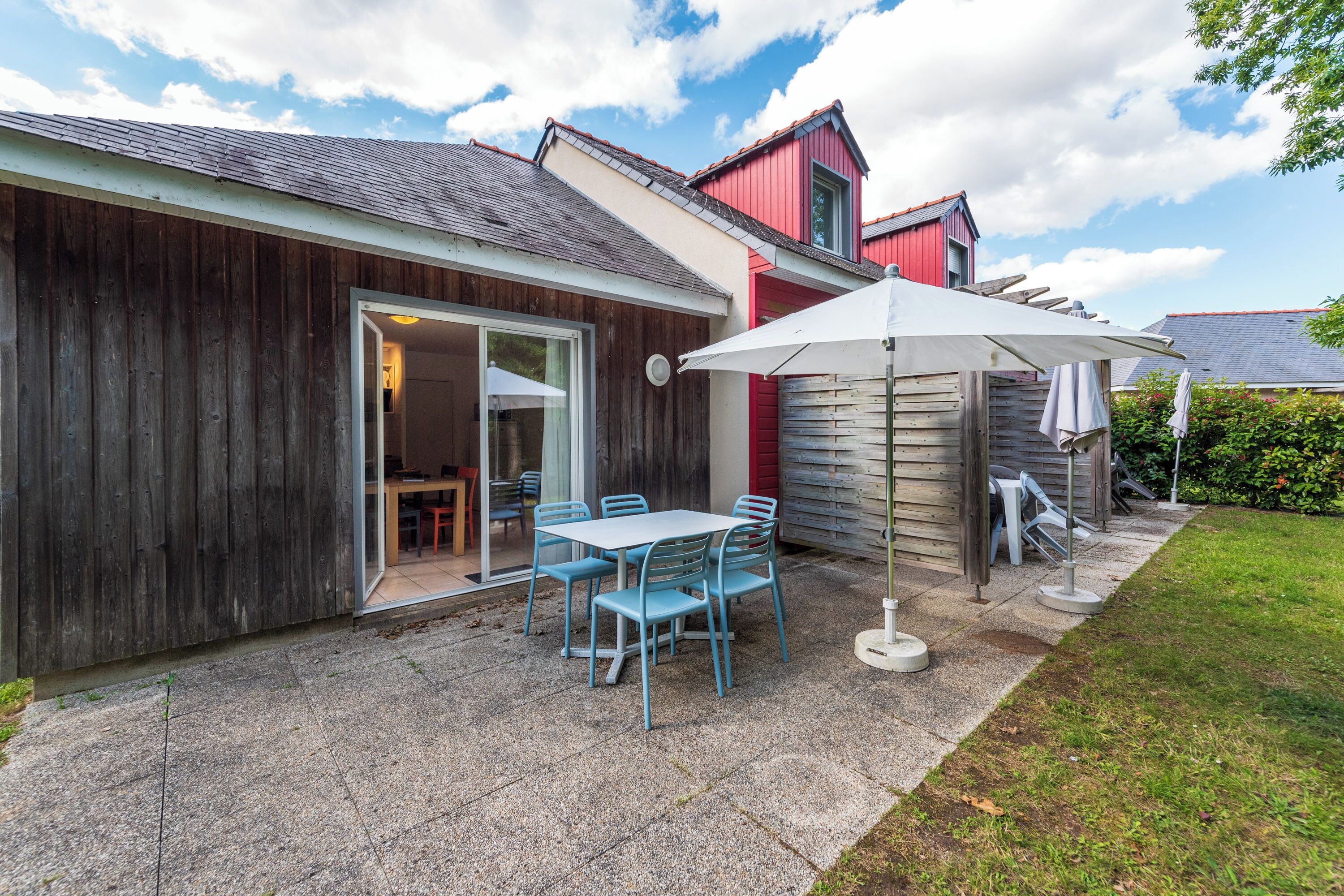 A patio with a table and chairs, a covered area with an umbrella, and a wooden building with a red door.