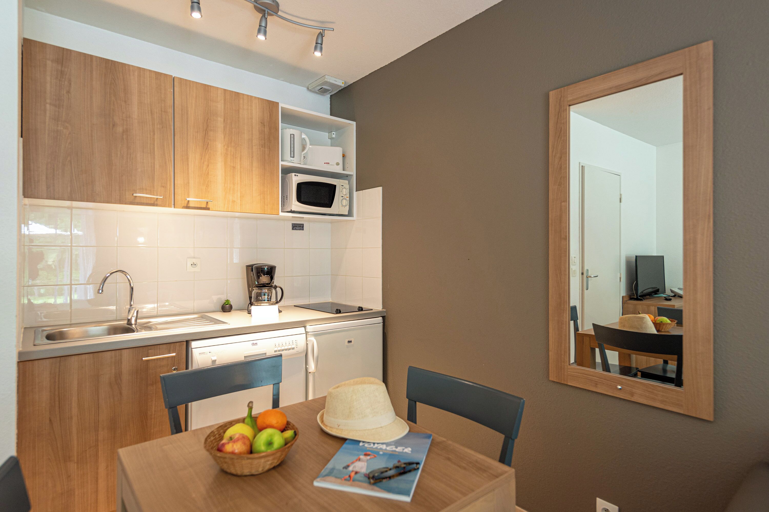 Une cuisine compacte avec des meubles en bois, un four à micro-ondes et une machine à café. On y trouve une table à manger avec des chaises et un panier de fruits.