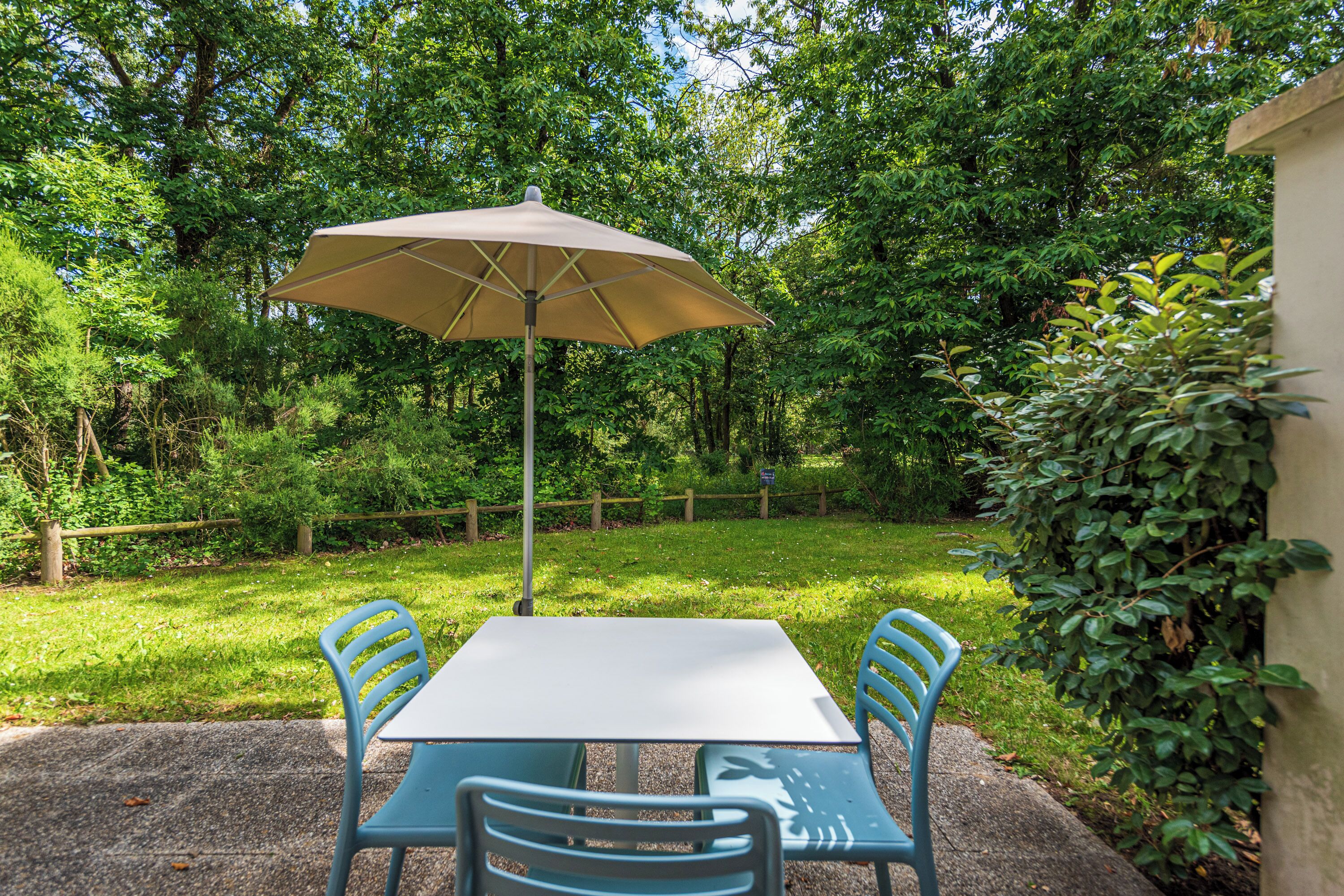 Un patio al aire libre con una mesa y sillas, una sombrilla color beige y abundante vegetación de fondo.