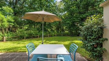 Un patio al aire libre con una mesa y sillas, una sombrilla color beige y abundante vegetación de fondo.