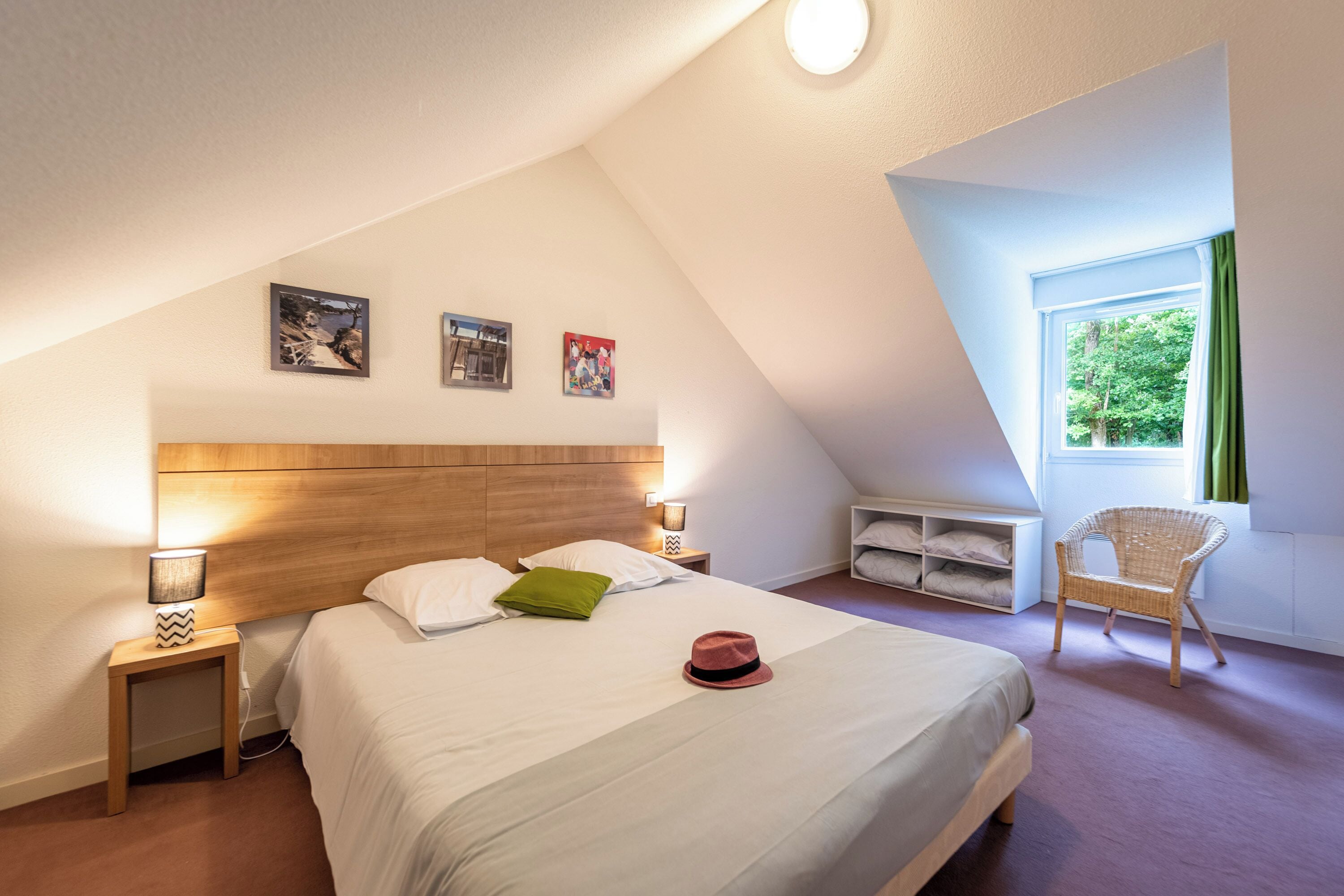 A bedroom with a sloped ceiling, a bed with white bedding, a green pillow, a red hat, a wicker chair, and a window with curtains.
