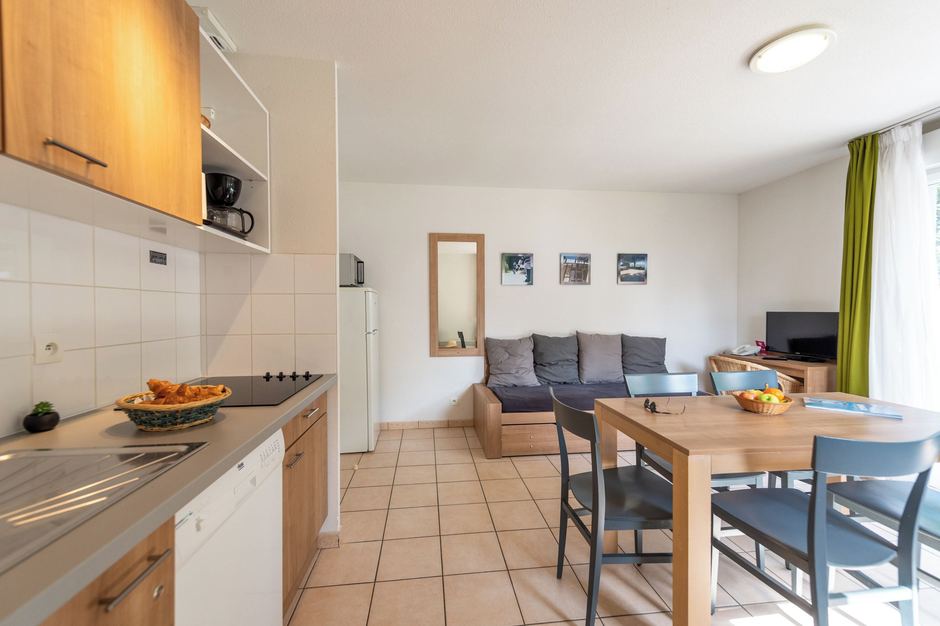 Cocina compacta con estufa, pileta y alacenas. Una mesa de comedor con sillas está en el centro. Un sofá y un televisor se ven al fondo.