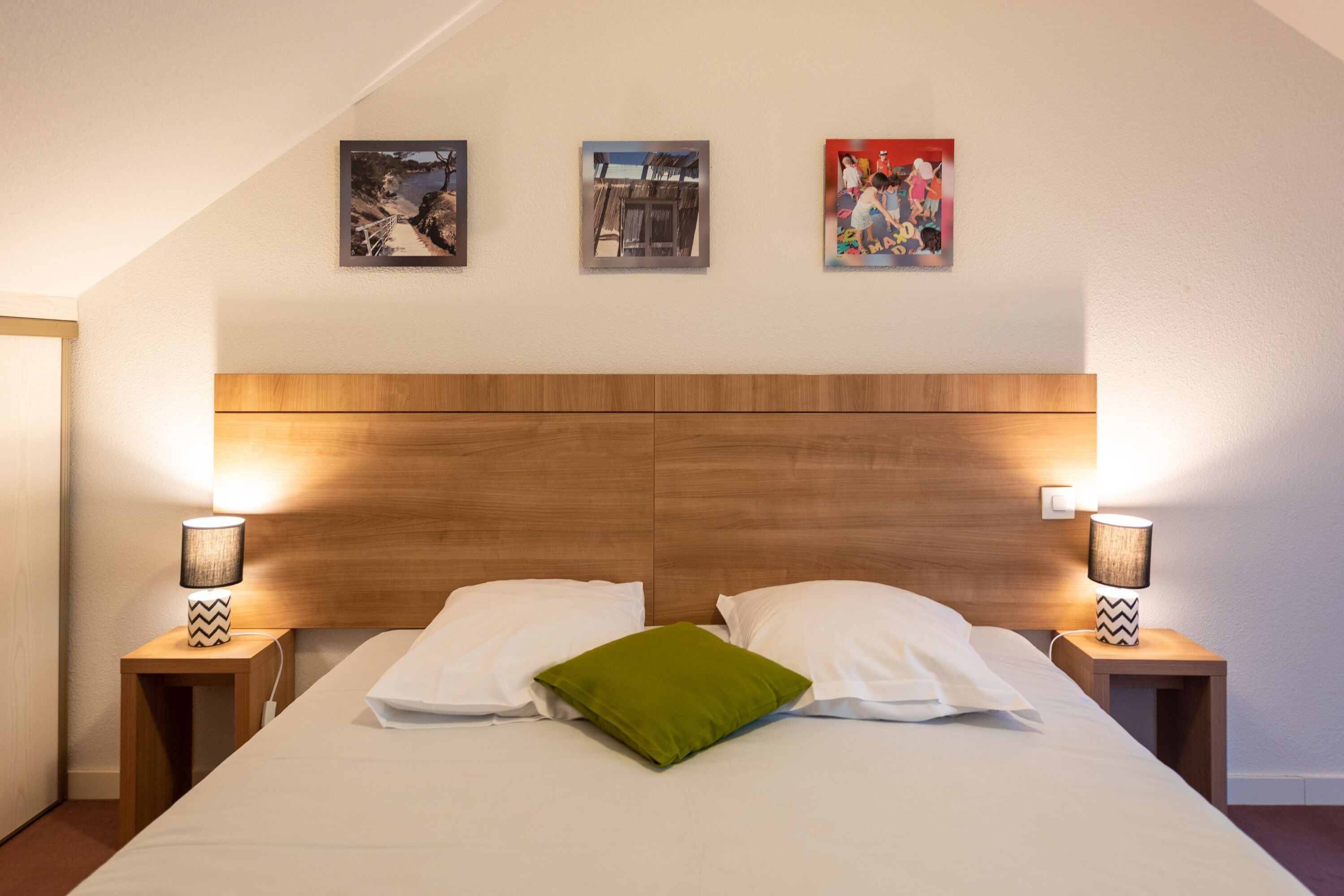 A neatly made bed with white linens and a green pillow, flanked by two bedside tables with lamps, in a room with a wooden headboard and three framed pictures on the wall.