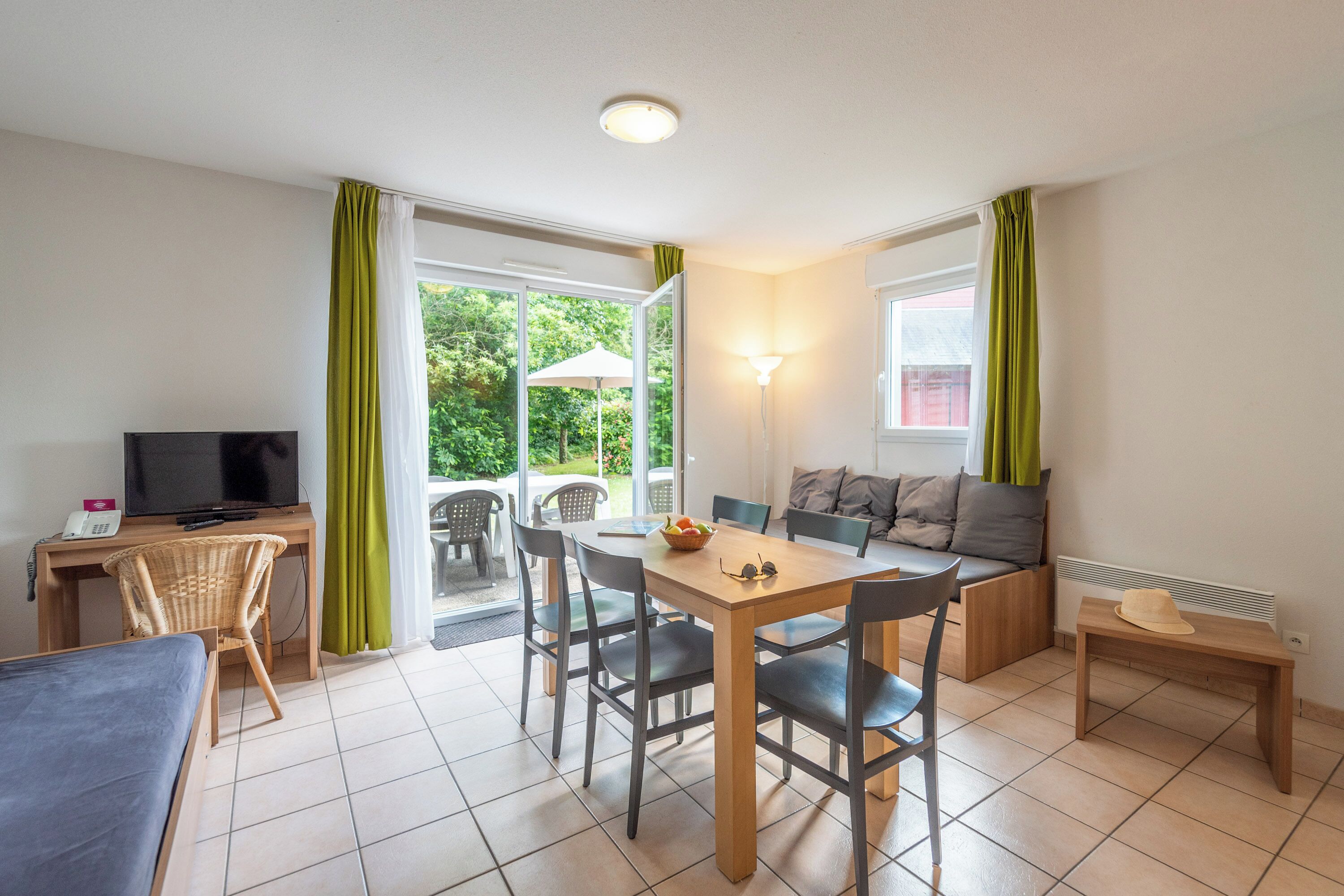 A room with a bed, a desk with a computer, a dining table with chairs, a sofa, and a small table with a hat.
