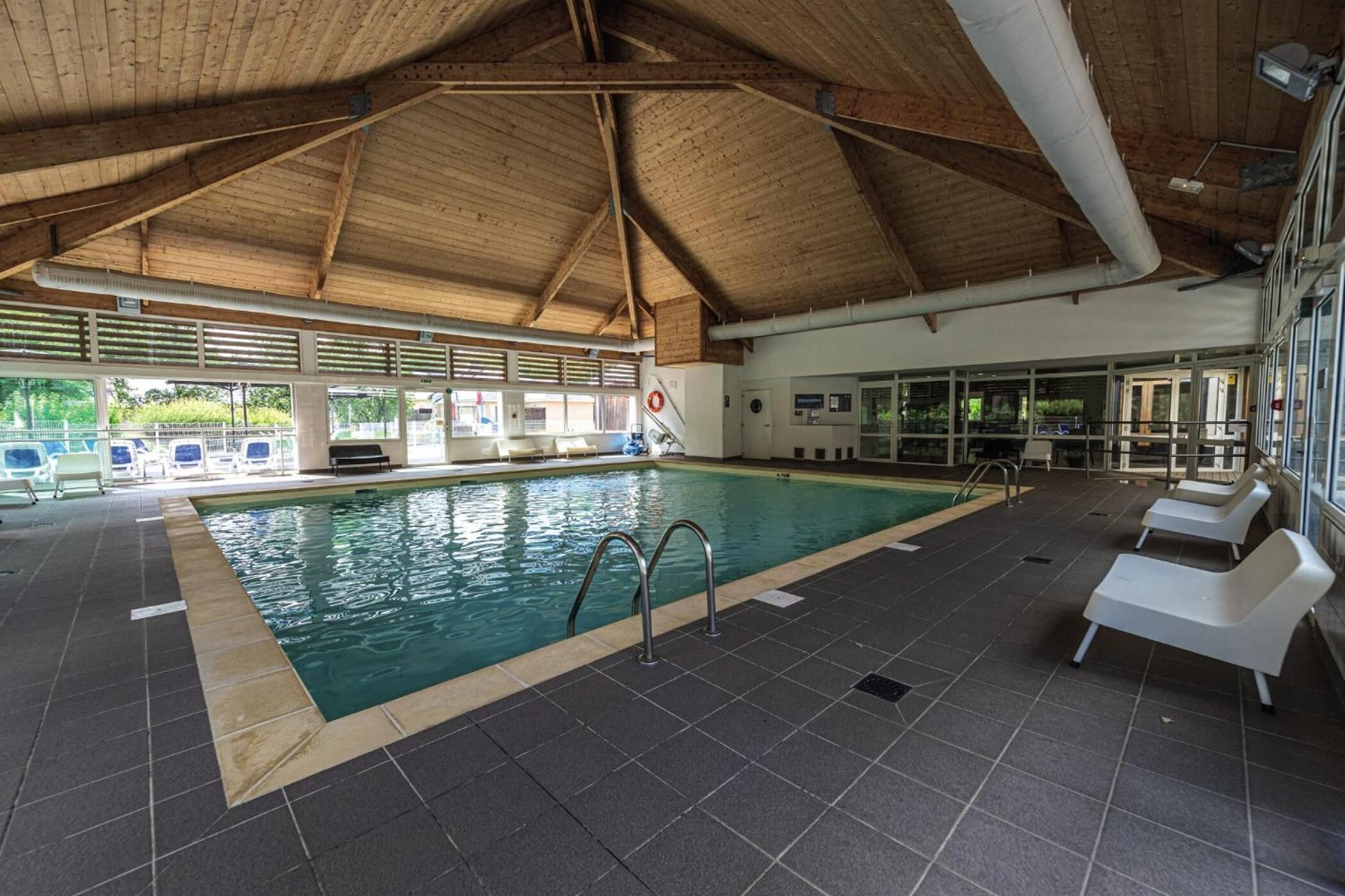 An indoor swimming pool with a tiled floor, a wooden ceiling, and a view of the outdoors through large windows.