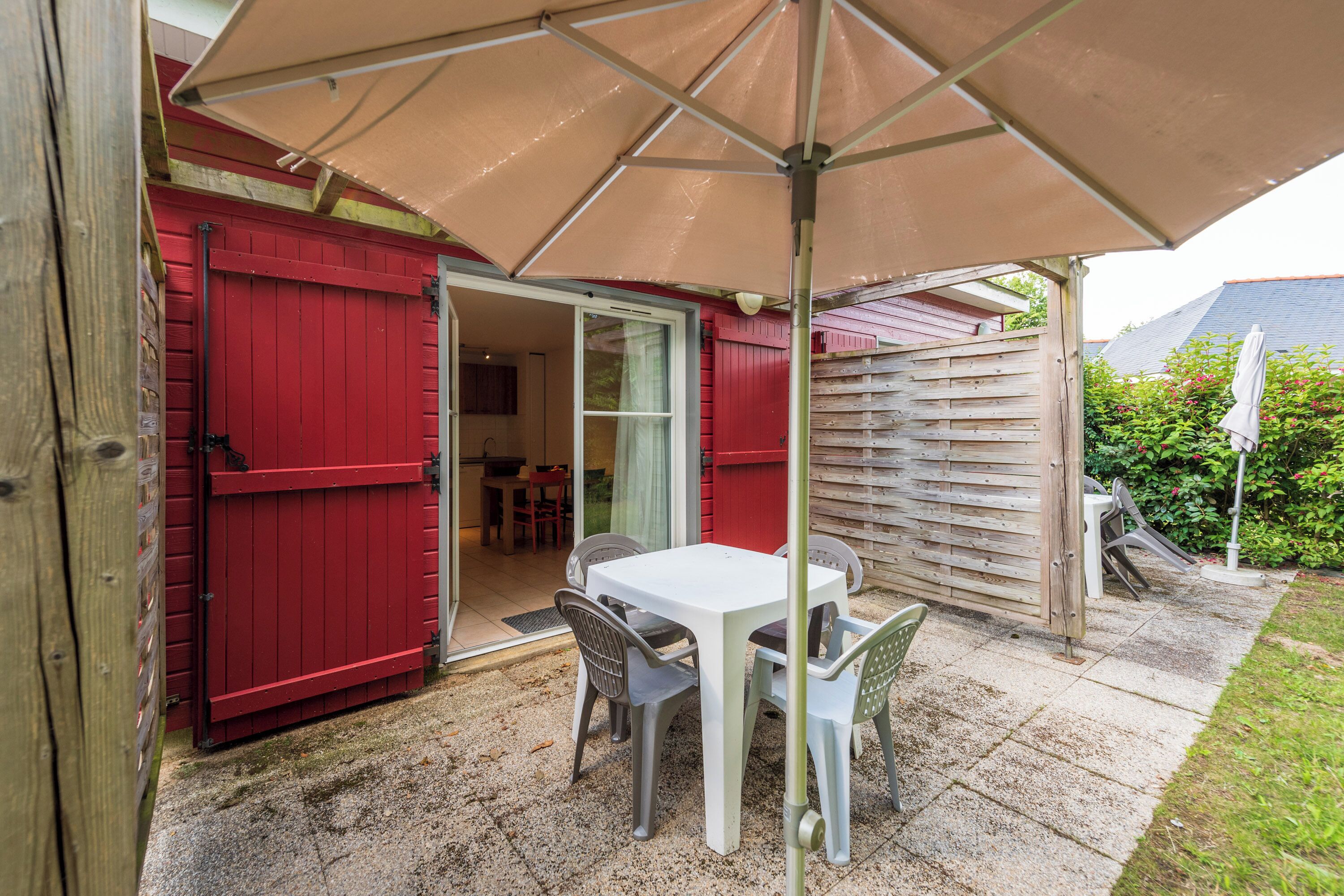 Un patio al aire libre con una mesa y sillas bajo un paraguas grande, una puerta roja y una cerca de madera.