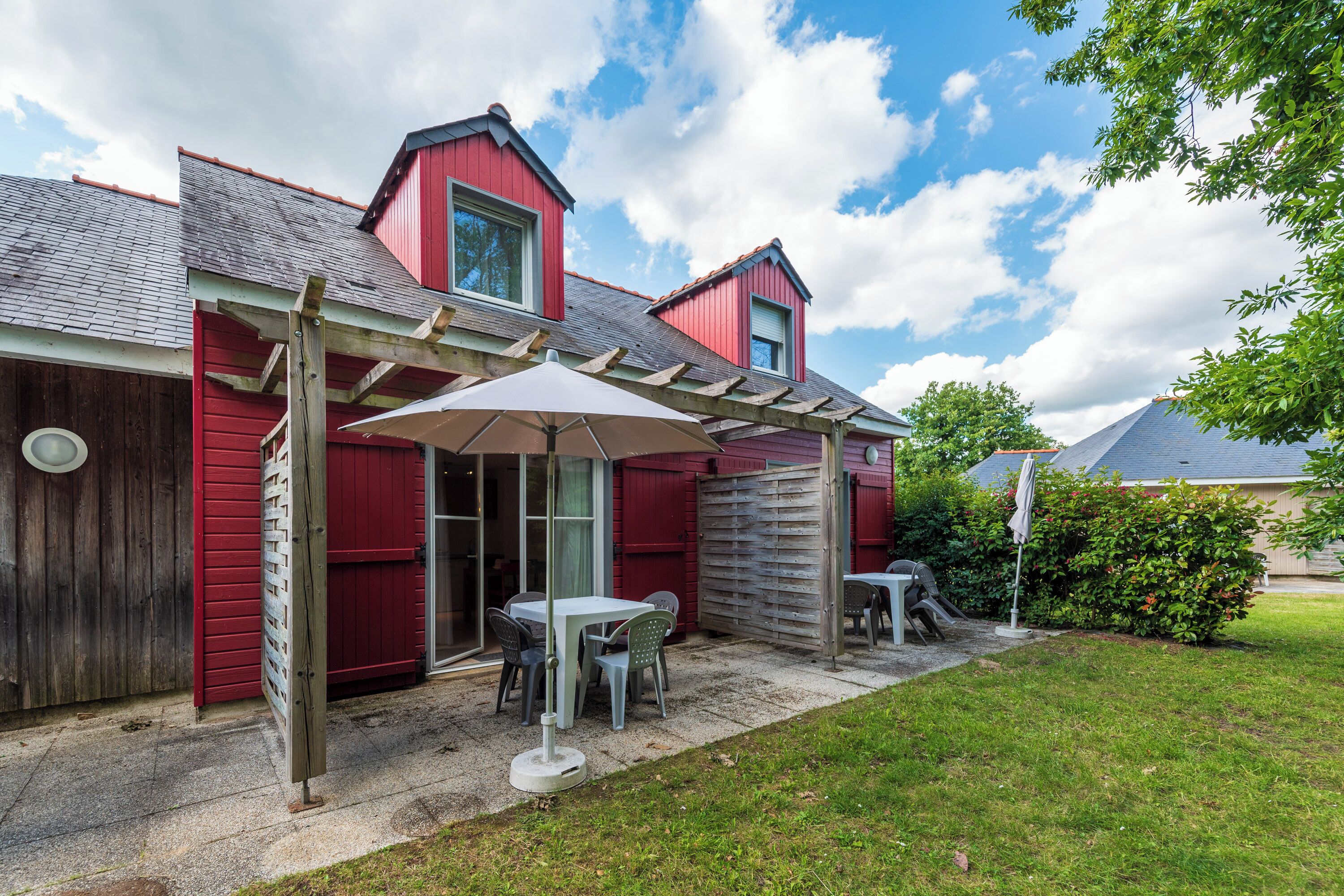 A red building with a covered patio area, a wooden pergola, and a small table with chairs.