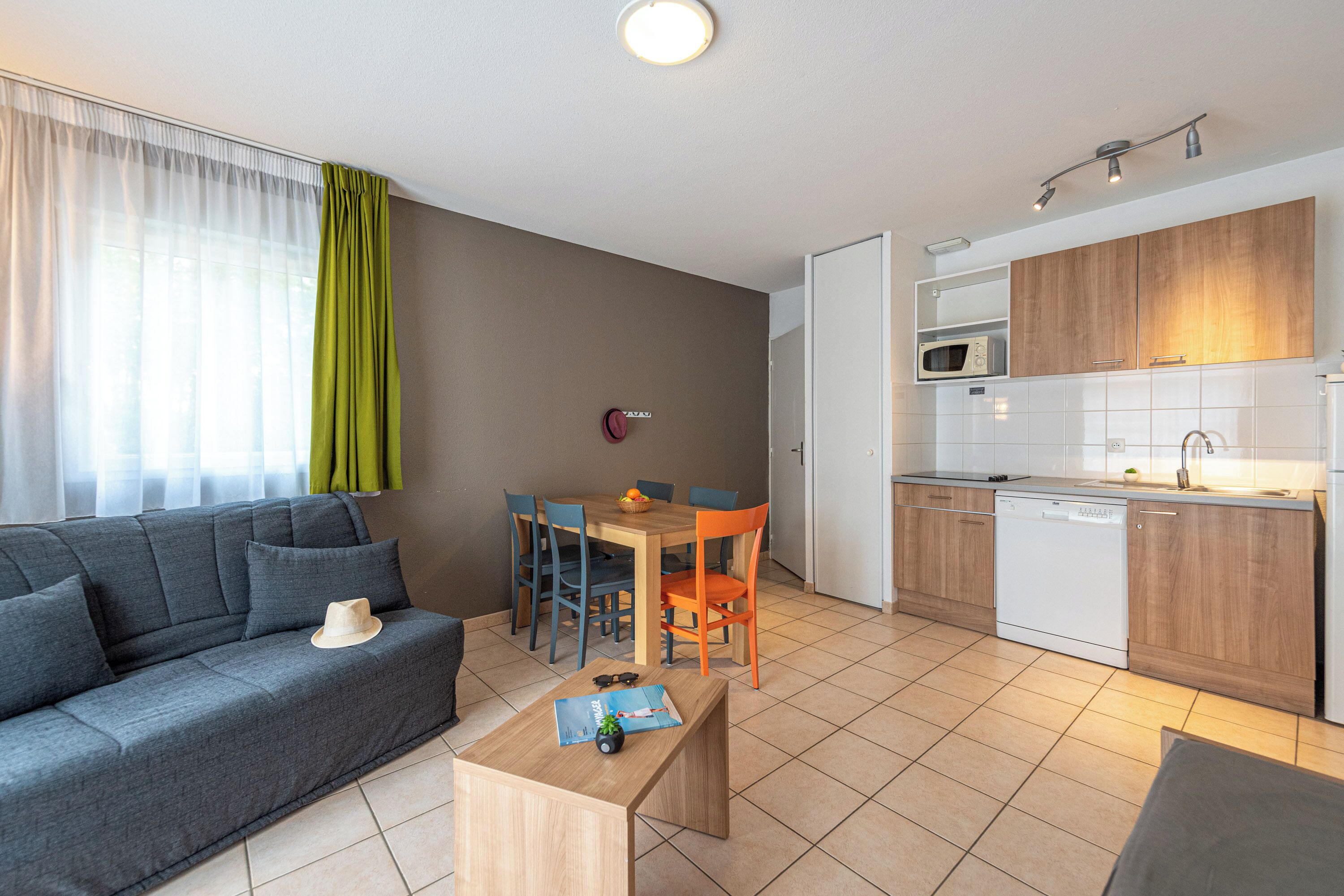 A modern living room with a sofa, a dining table, and chairs. There is a kitchen area with cabinets and a dishwasher.