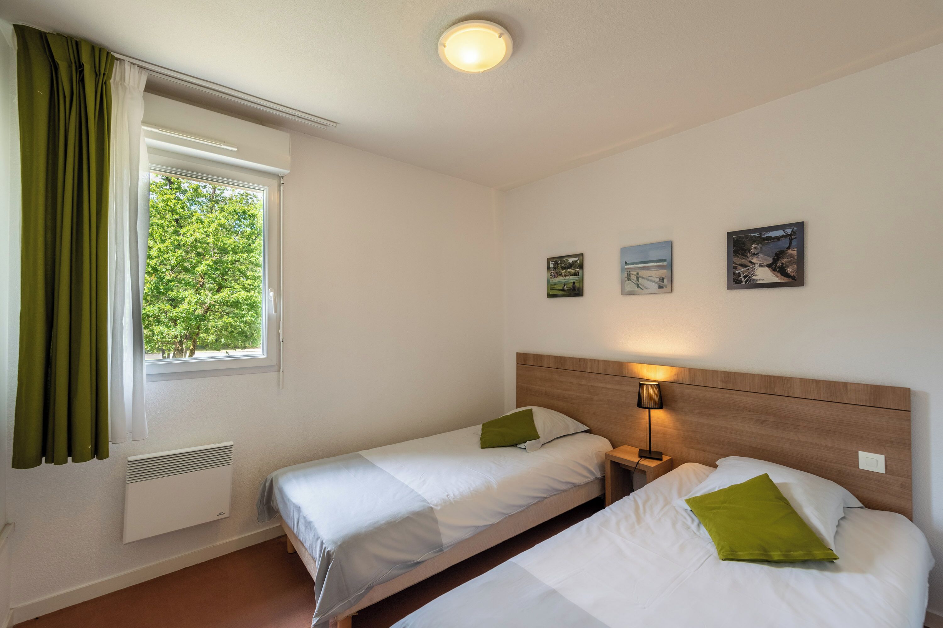 A room with two beds, a window with curtains, a wall-mounted heater, and three framed pictures.