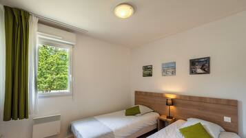A room with two beds, a window with curtains, a wall-mounted heater, and three framed pictures.