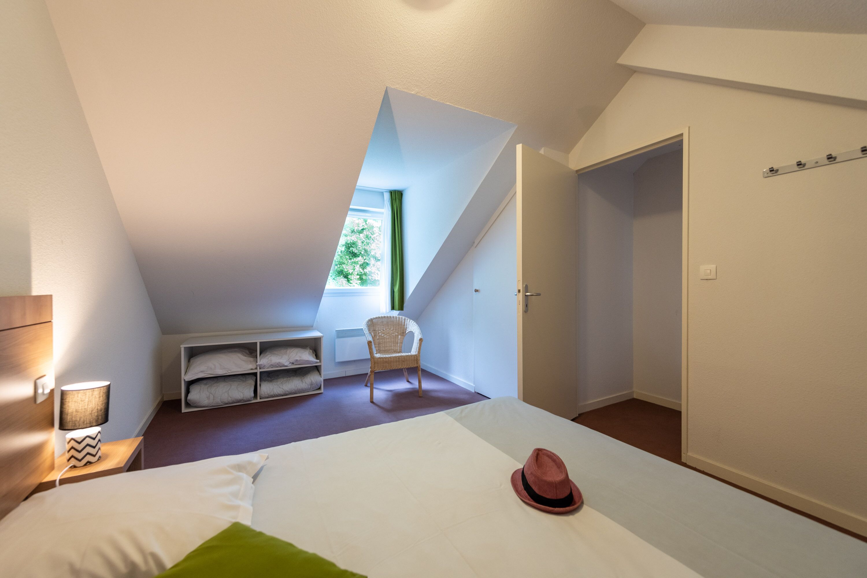 A bedroom with a sloped ceiling, a bed, a chair, a nightstand, a lamp, a window, and a door.