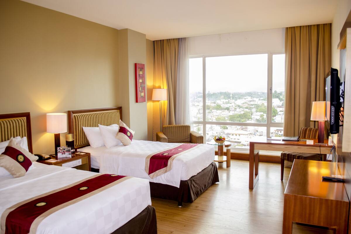 Habitación de hotel con dos camas, un escritorio, una silla, un televisor y un amplio ventanal con vistas a la ciudad.