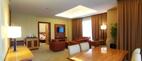 A hotel room with a wooden dining table, two sofas, a TV, and a plant.