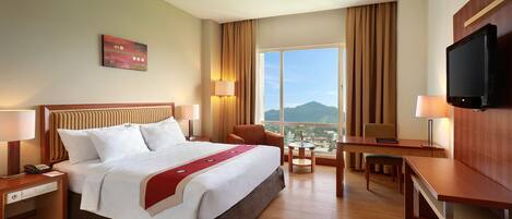 A hotel room with a large bed, a desk, a chair, a TV, and a view of a cityscape through large windows.