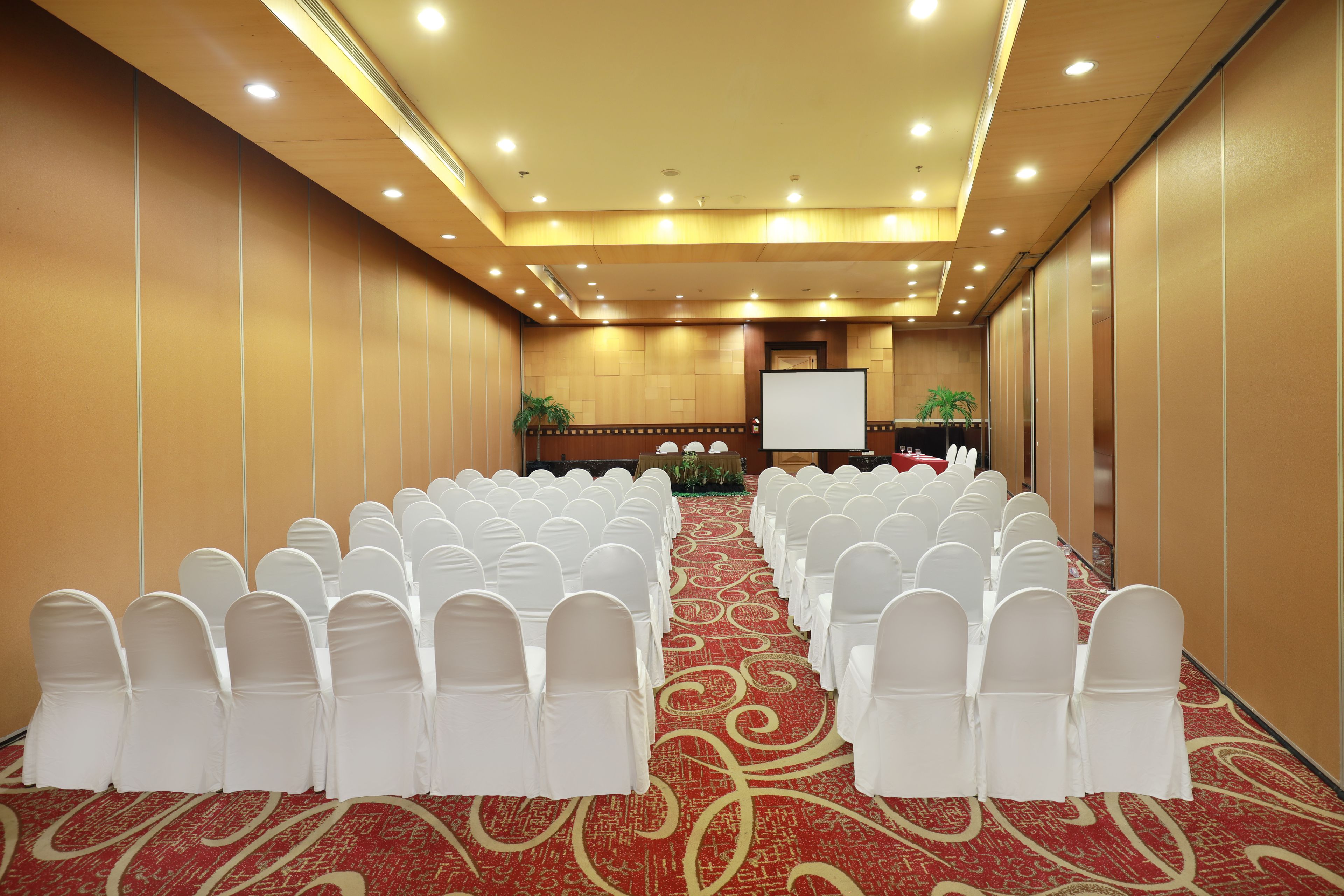 A conference room with rows of white chairs, a red carpet, and a projector screen at the front.