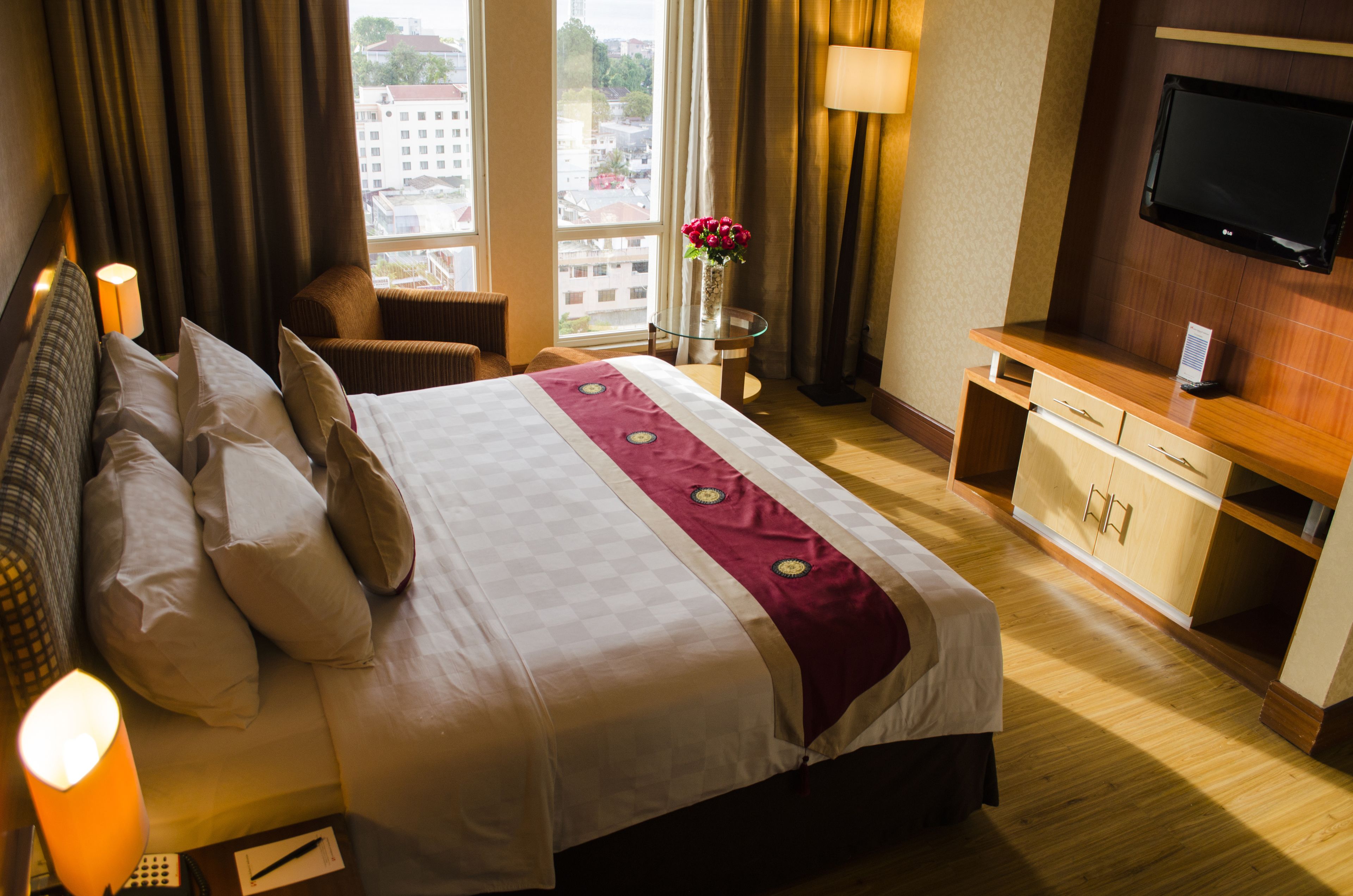 A hotel room with a large bed, a desk with a flat-screen TV, a window with a view of buildings, and a vase with flowers on the desk.