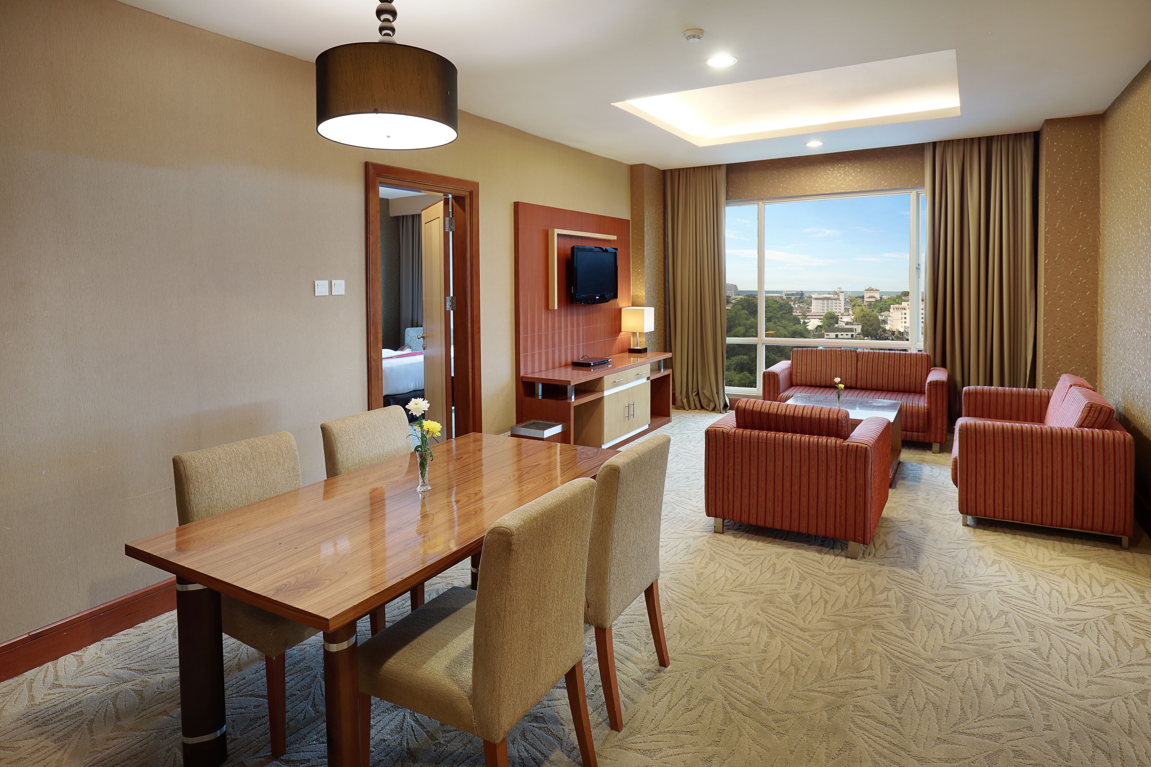 A hotel room with a dining table, chairs, a sofa set, a TV, and a large window with a city view.