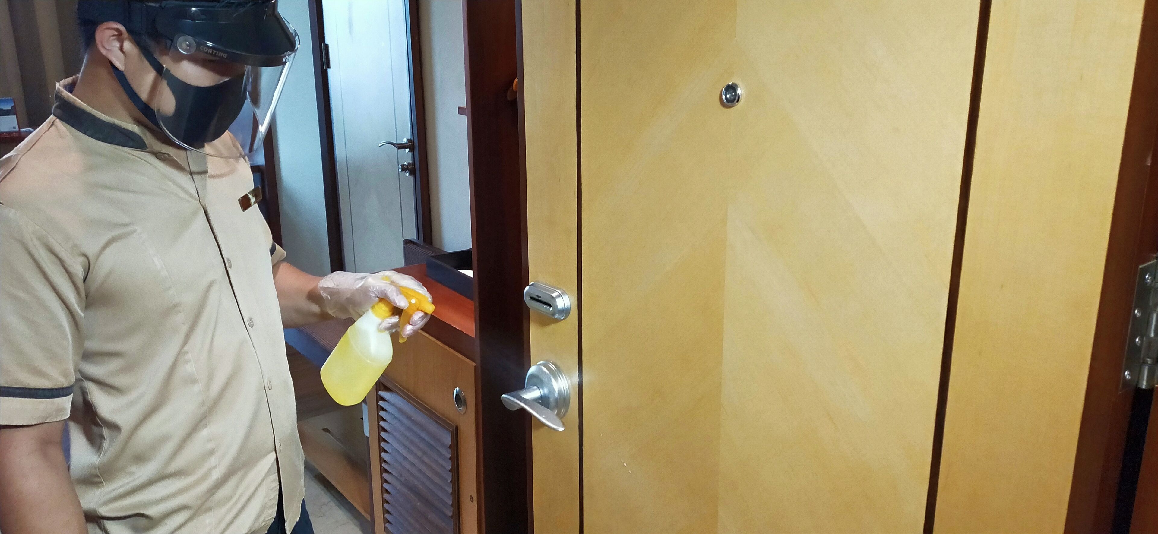 A person in a uniform holding a spray bottle, standing in front of a wooden door with a metal handle and a keyhole.