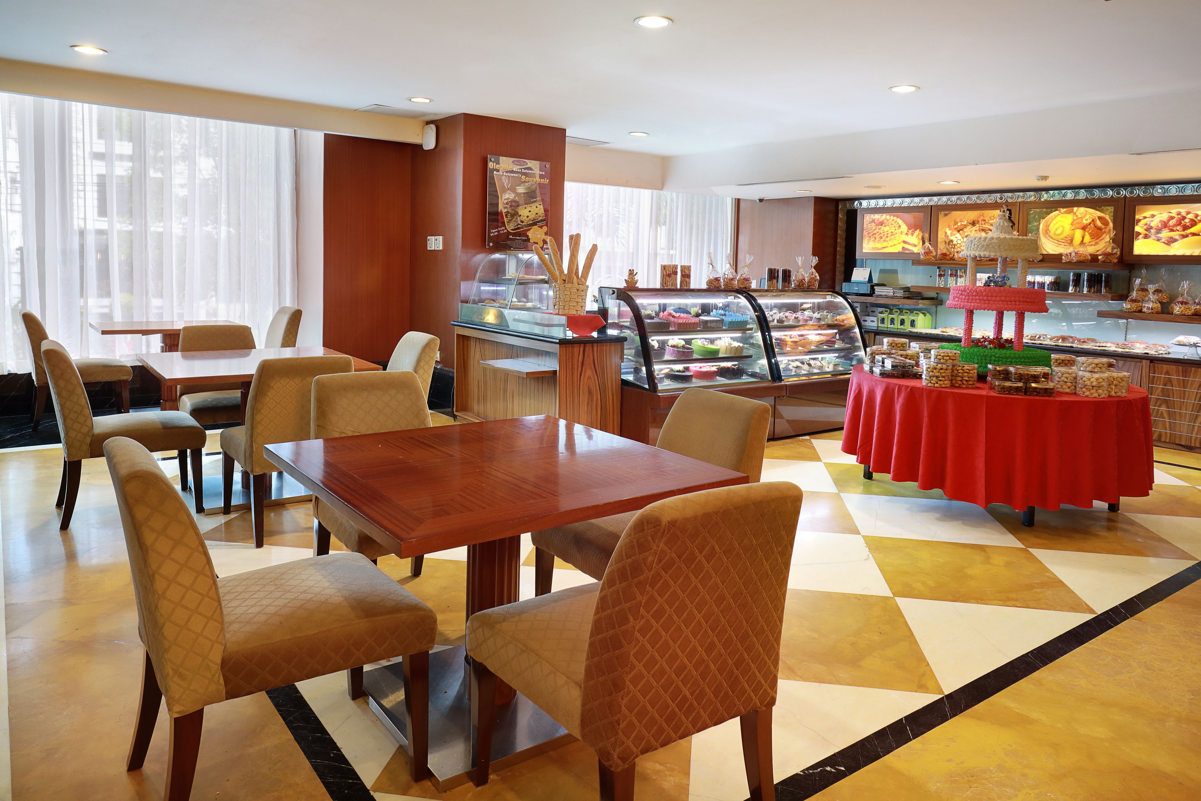 Uma área de refeições com mesa e cadeiras de madeira, uma vitrine de sobremesas e um buffet com toalha de mesa vermelha.
