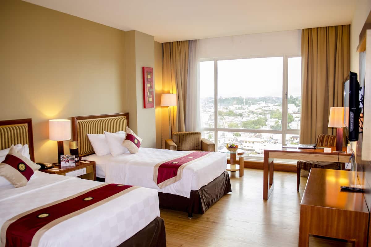 Habitación de hotel con dos camas, un escritorio, una silla, un televisor y un ventanal con vistas a la ciudad.