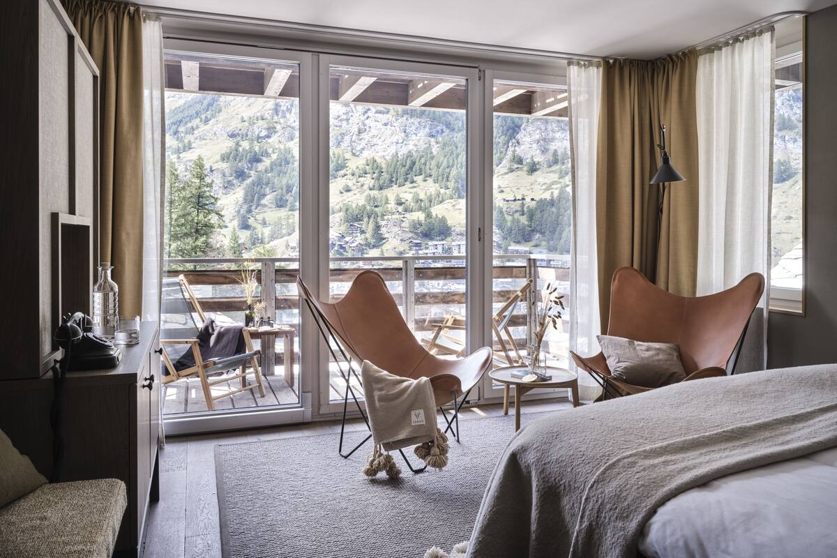 Quarto de hotel com uma grande porta de vidro deslizante que dá para uma varanda, duas poltronas de couro, uma pequena mesa redonda e uma cama com manta branca.
