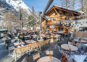 A cozy outdoor seating area with tables and chairs, surrounded by wooden buildings and snow-covered mountains. - CERVO Mountain Resort (Zermatt)
