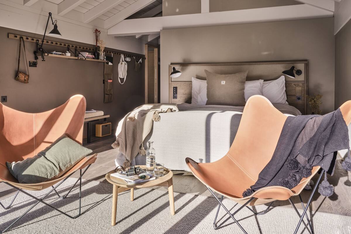 Quarto com cama grande, duas poltronas, uma mesinha e prateleira de madeira com objetos pessoais.