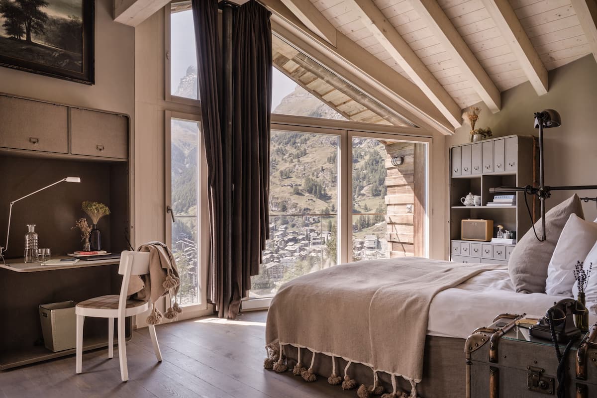 Quarto com uma cama grande, uma escrivaninha com cadeira, uma janela com cortinas e vista para uma paisagem montanhosa.