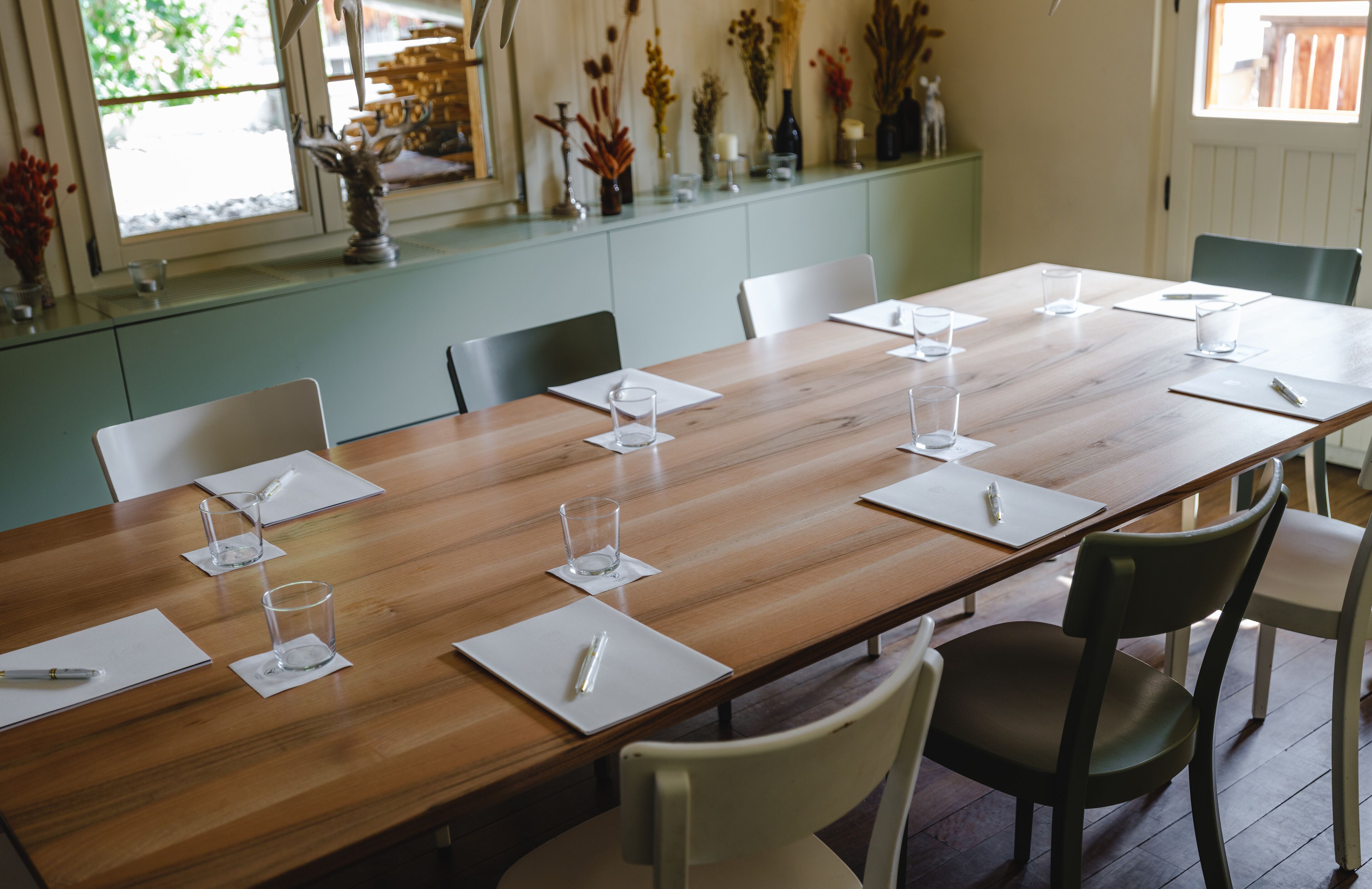 Un salón de conferencias con una larga mesa de madera, sillas blancas y vasos de vidrio vacíos sobre posavasos.