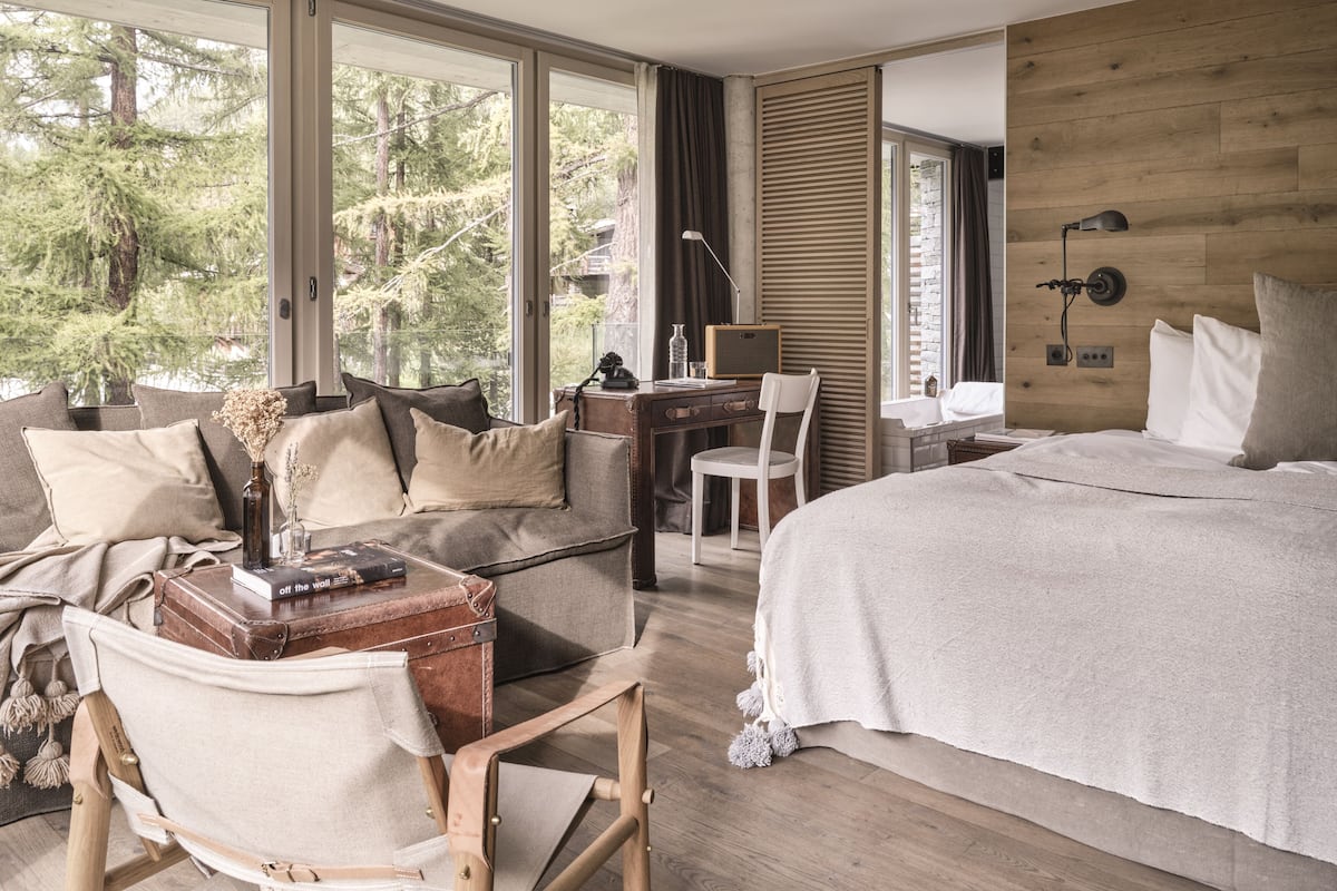 Quarto moderno com uma cama grande, uma escrivaninha e um sofá. Janelas amplas oferecem vista para árvores.