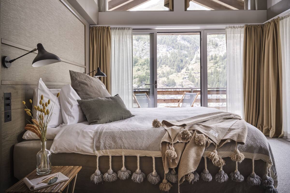 Quarto com uma cama grande, mesa de cabeceira com abajur, janela com cortinas e vista para uma paisagem montanhosa.