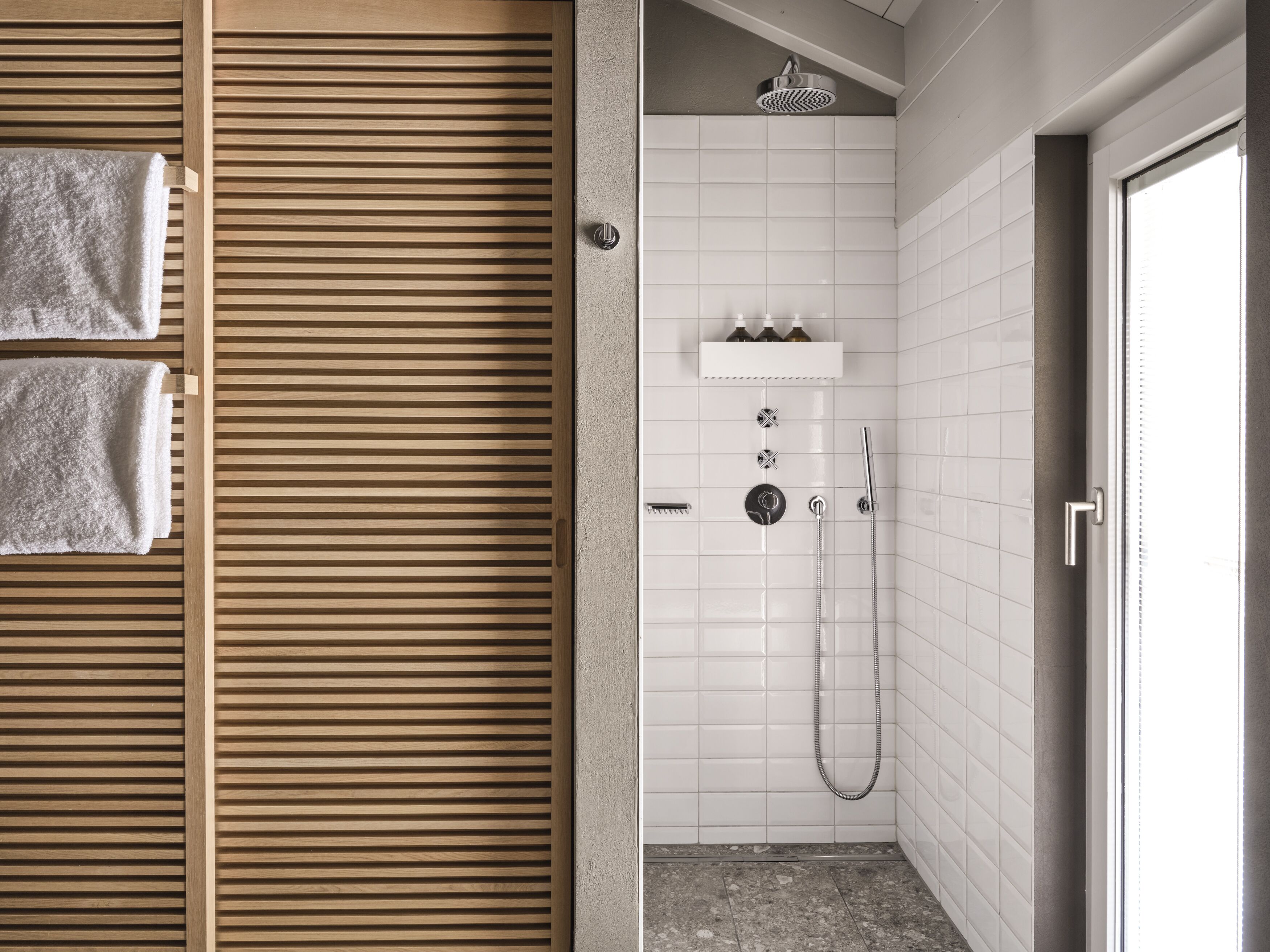 Et moderne badeværelse med dør i trælameller, hvide flisevægge, et bruseområde med håndholdt brusehoved og en lille hylde med toiletartikler.