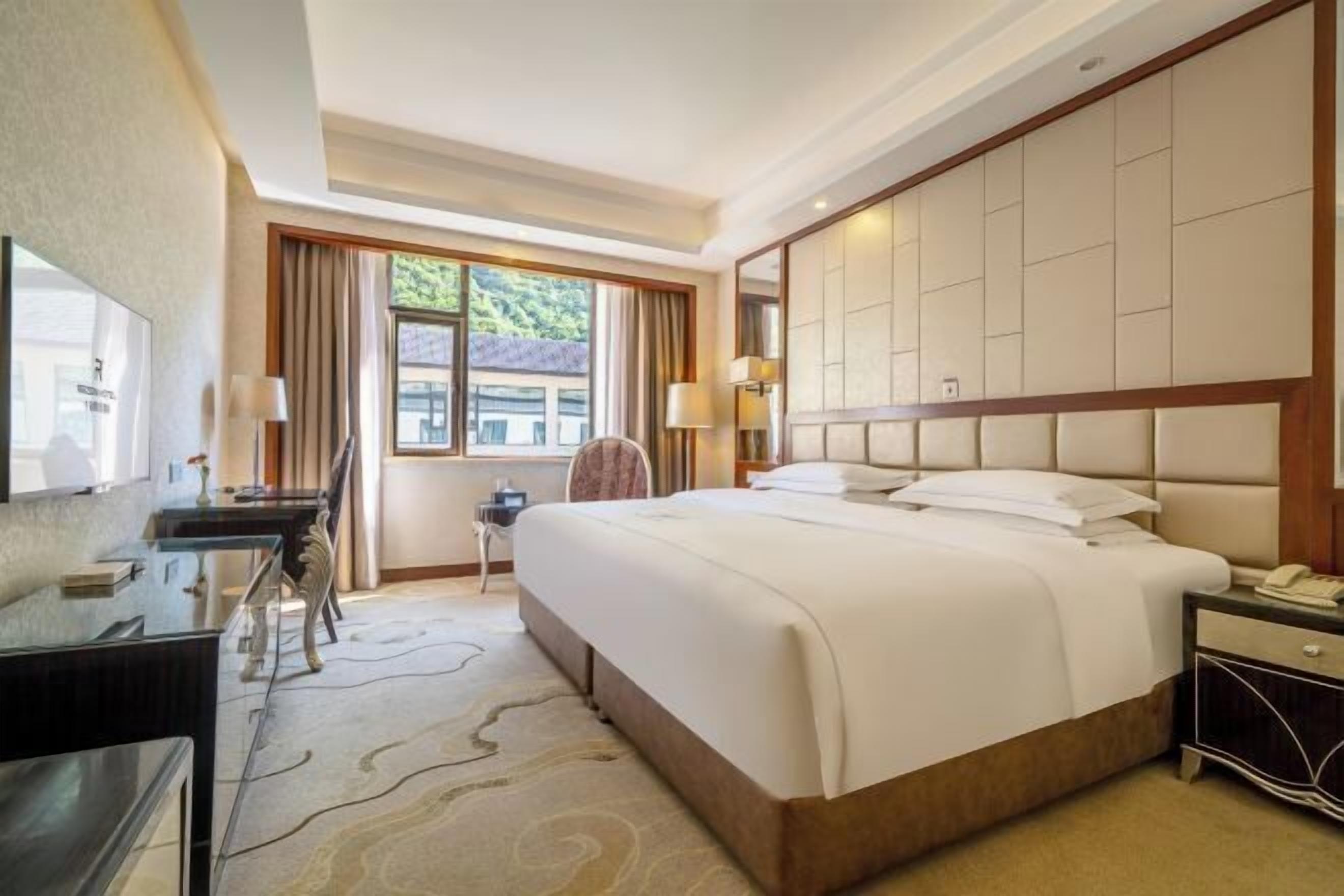 Habitación de hotel con una cama grande, un escritorio, una silla, un televisor y una ventana con cortinas.