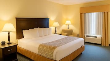 A hotel room with a large bed, two bedside tables with lamps, a chair, a window with curtains, and an air conditioning unit.