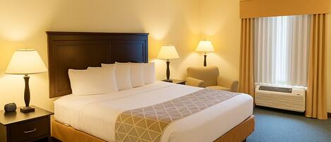 Una habitación de hotel con una cama grande, dos mesitas de noche con lámparas, una silla, una ventana con cortinas y un aire acondicionado.