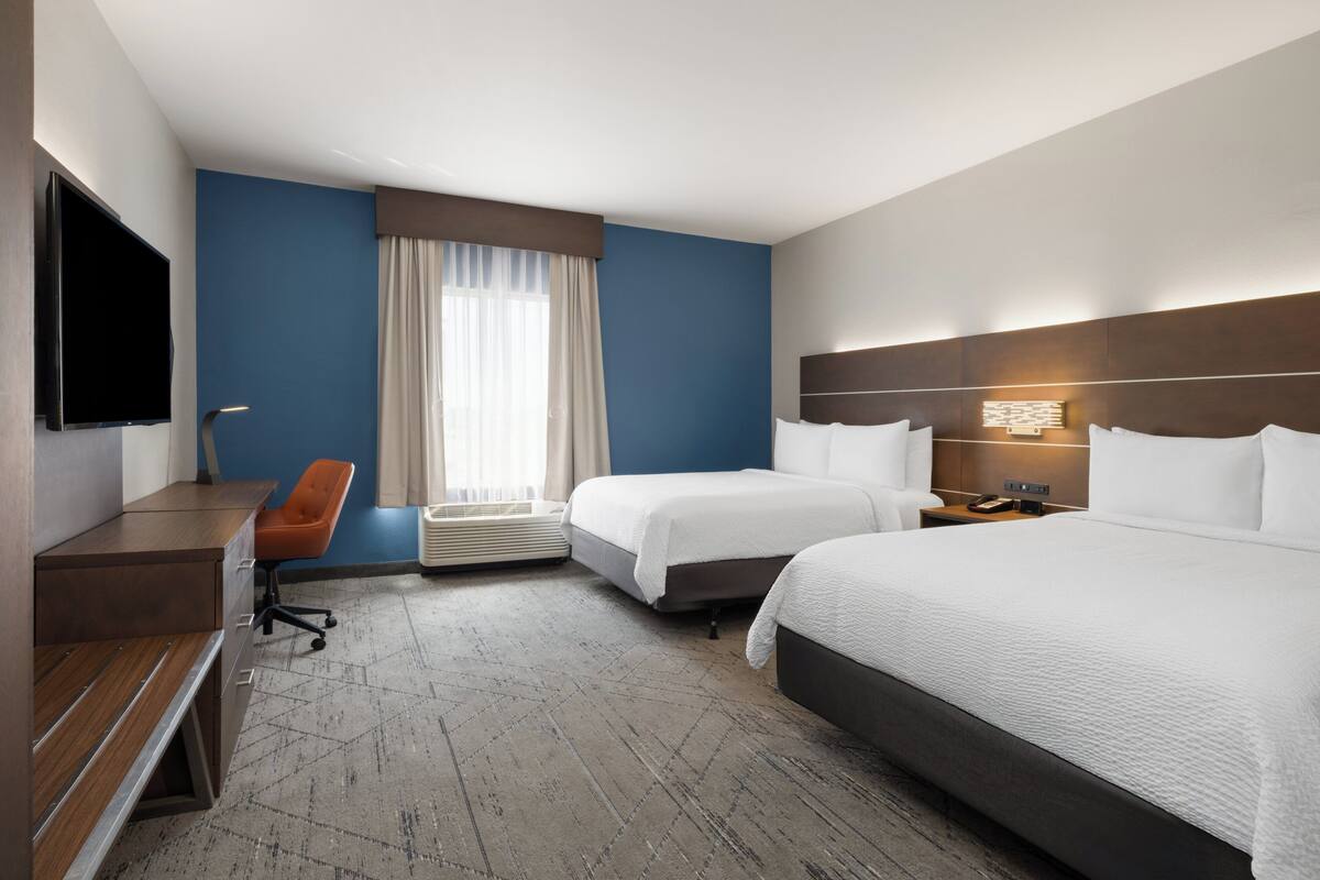 Ein Hotelzimmer mit zwei Betten, einem Schreibtisch, einem Stuhl, einem Flachbildfernseher und einem Fenster mit Vorhängen.