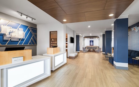 A modern hotel reception area with a wooden reception desk, a seating area with a table and chairs, and a large wall with geometric patterns.