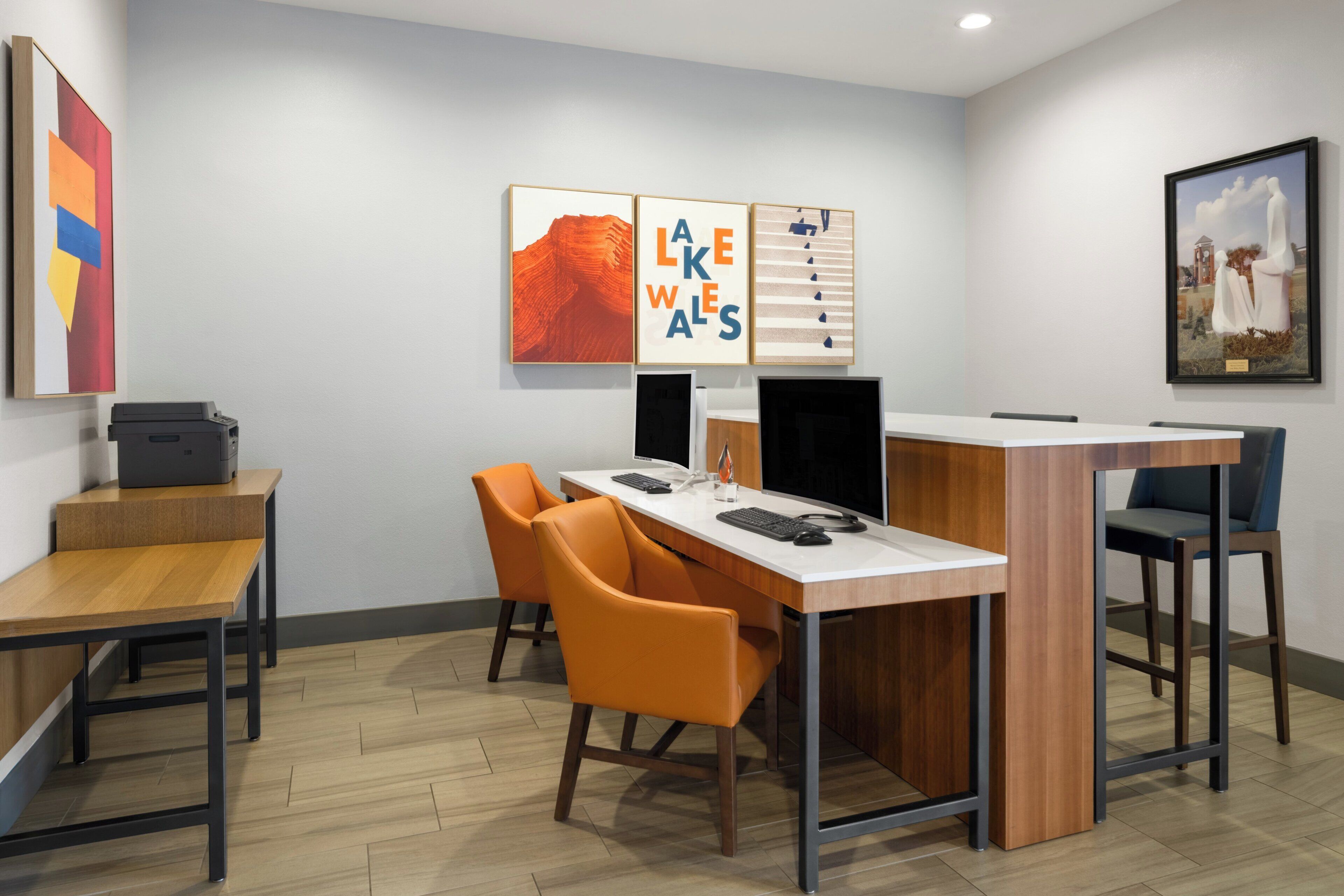 Un espace de bureau moderne avec un bureau en bois, deux chaises orange, un ordinateur et des œuvres d’art encadrées accrochées au mur.