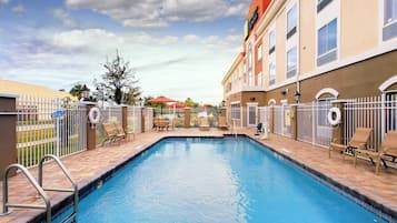 Una piscina con valla blanca, sillas de descanso y un edificio con un letrero.